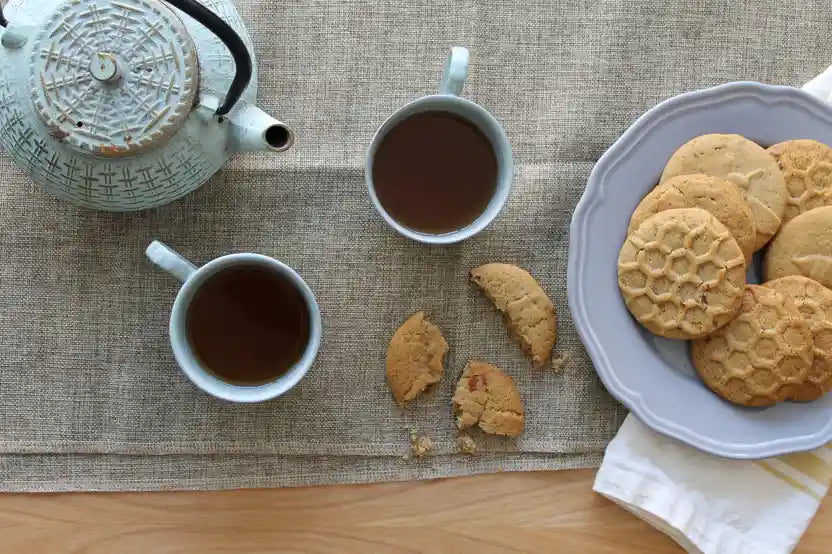 Two cups of tea with a teapot and plate of cookies on a textured surface.
