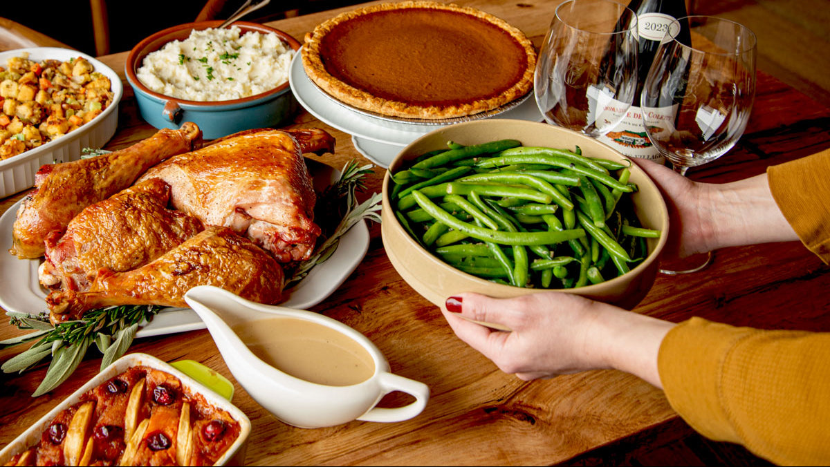 hands placing a plate of green beans on a  table set for Thanksgiving with turkey, pumpkin pie, gravy and wine