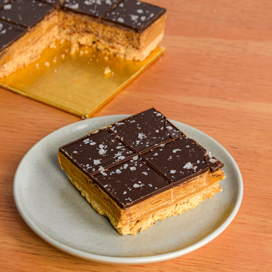 A square slice of chocolate sqaure with chocolate top, caramel middle layer, and biscuit base, served on a light gray plate on a wooden table. A larger cut piece of the same dessert is visible in the background on a gold board.