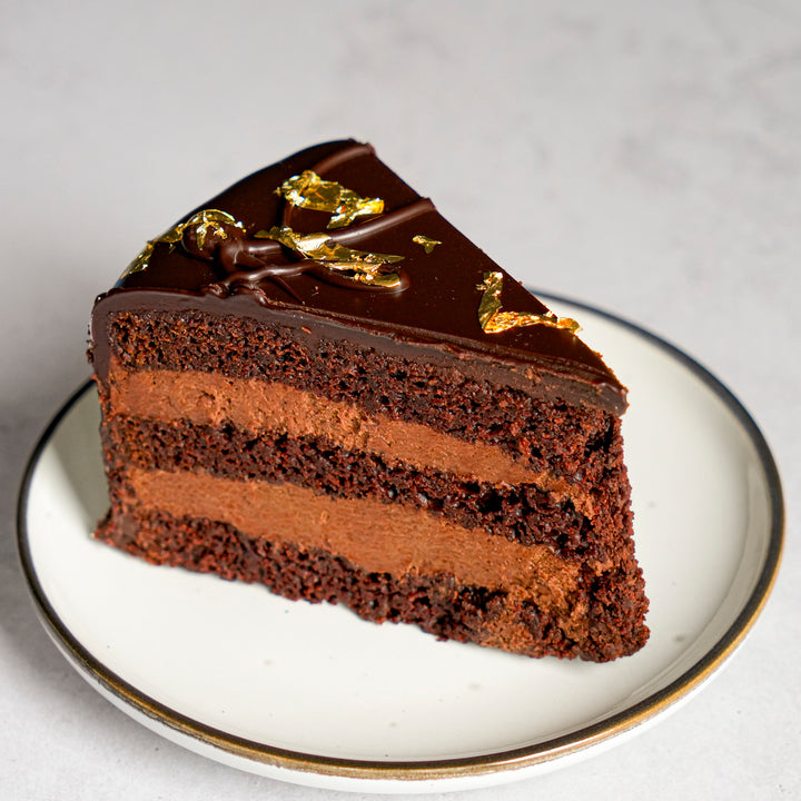 A triangular slice of chocolate cake with two layers of chocolate cream filling and a glossy chocolate glaze on top, decorated with gold leaf flakes, served on a white plate with a gold rim.