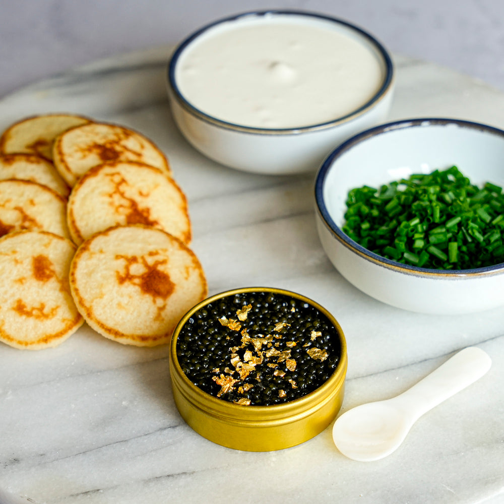 A gourmet food presentation on a white marble board featuring a gold tin of black caviar with gold flakes, a small white spoon, a stack of golden-brown mini pancakes, a bowl of creamy white sauce, and a bowl of chopped green chives.