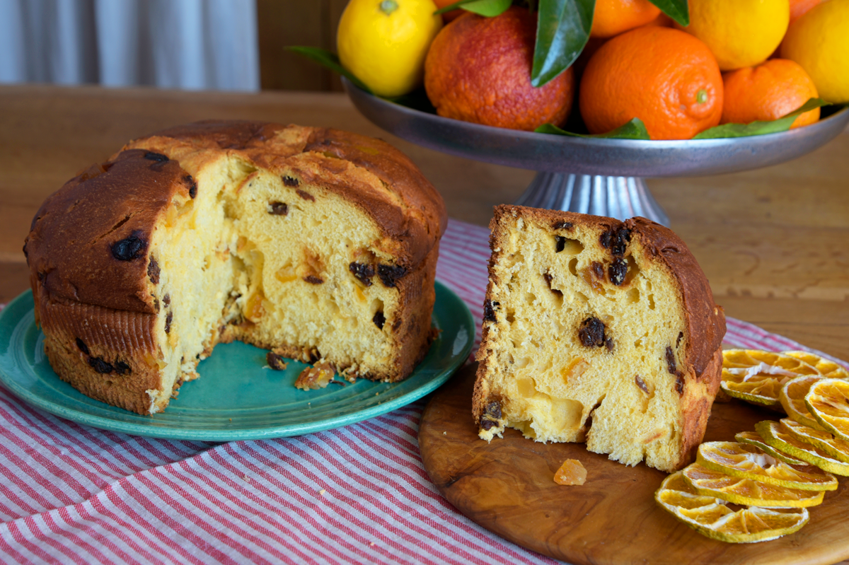 Panettone cake with a slice cut out, placed on a teal plate with a wooden board beside it, against a background of fruits.