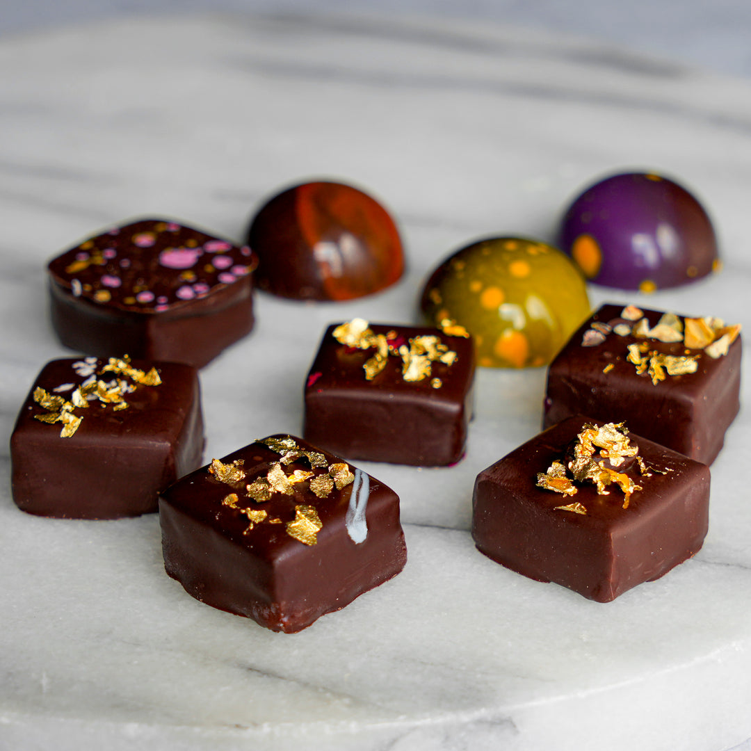An assortment of gourmet chocolate domes and square truffles decorated with gold leaf flakes, arranged on a white marble surface.