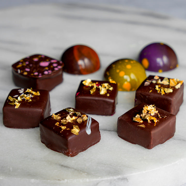 An assortment of gourmet chocolate domes and square truffles decorated with gold leaf flakes, arranged on a white marble surface.
