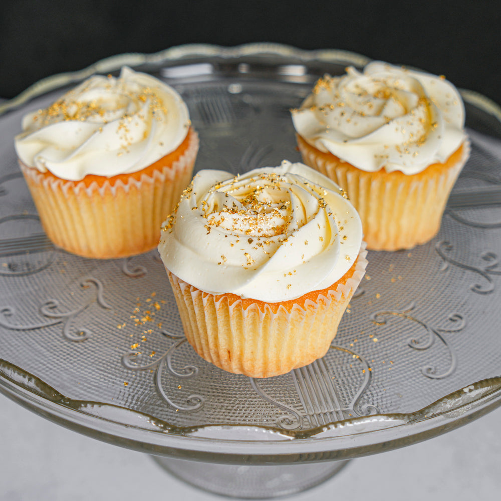 Three frosted cupcakes in paper liners on a clear glass cake stand, topped with white frosting and a sprinkle of golden crumbs, against a dark background.