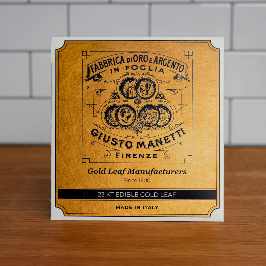 A square white card or box front with a large gold-toned label panel reading 'Fabbrica di Oro e Argento in Foglia' and 'Giusto Manetti Firenze,' standing on a wooden surface against a white tiled wall. Text also includes 'Gold Leaf Manufacturers,' '23 KT EDIBLE GOLD LEAF,' and 'Made in Italy.