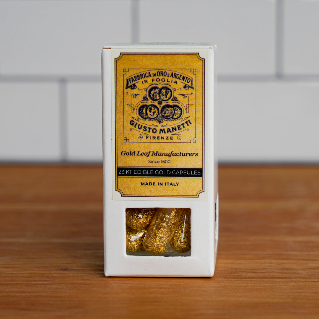 A white rectangular box of gold leaf capsules with a prominent yellow label reading 'Giusto Manetti Gold Leaf Manufacturers' and a clear window at the bottom showing the gold capsules inside, set on a wooden surface against a light gray tiled background.