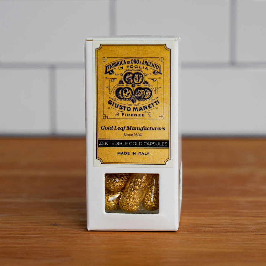 A white rectangular box of gold leaf capsules with a prominent yellow label reading 'Giusto Manetti Gold Leaf Manufacturers' and a clear window at the bottom showing the gold capsules inside, set on a wooden surface against a light gray tiled background.