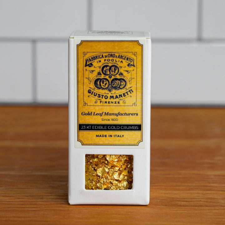 A white rectangular box of gold leaf crumbs with a large yellow front label reading 'Giusto Maretti' and a clear window showing metallic gold flakes inside, set on a wooden surface against a blurred white tiled background.