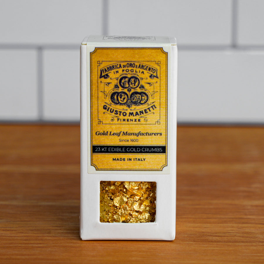 A white rectangular box of gold leaf crumbs with a large yellow front label reading 'Giusto Maretti' and a clear window showing metallic gold flakes inside, set on a wooden surface against a blurred white tiled background.