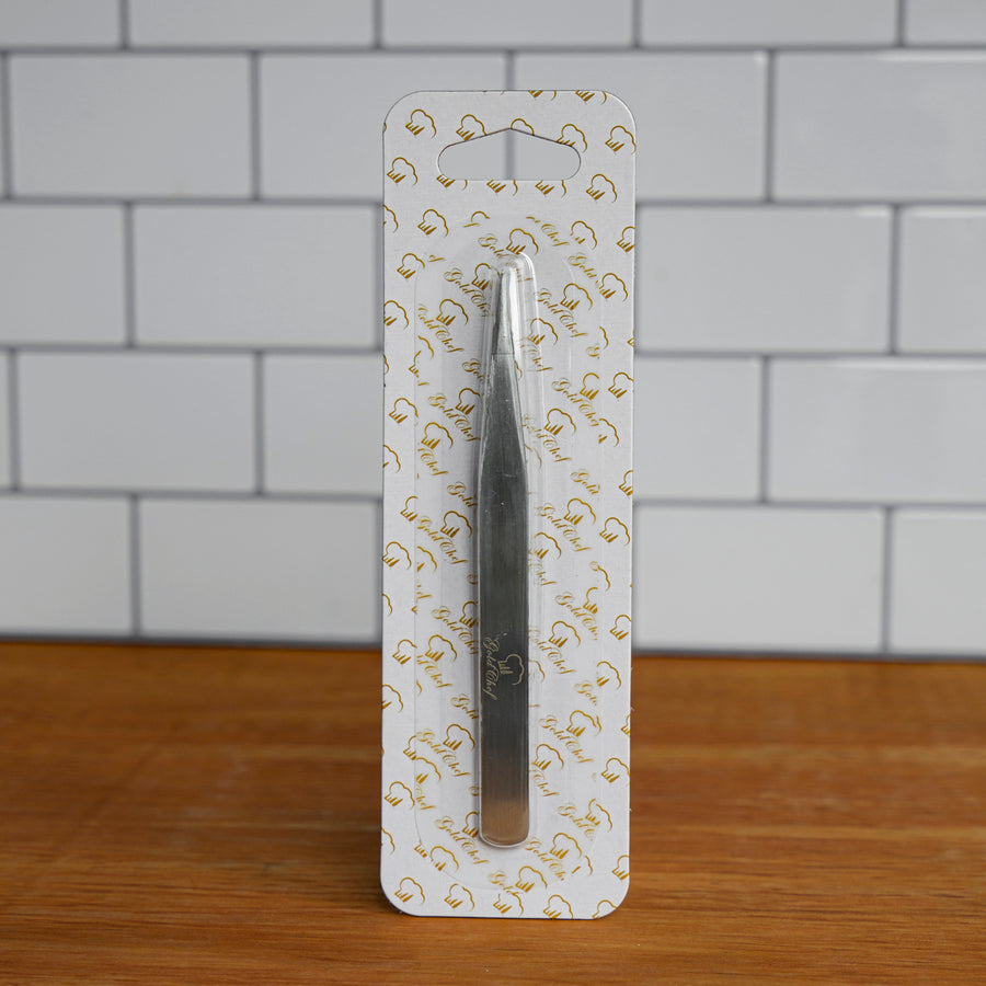 A pair of silver metal tweezers in clear plastic blister packaging with a white backing card printed with gold script, standing on a wooden surface against a white subway tile wall.