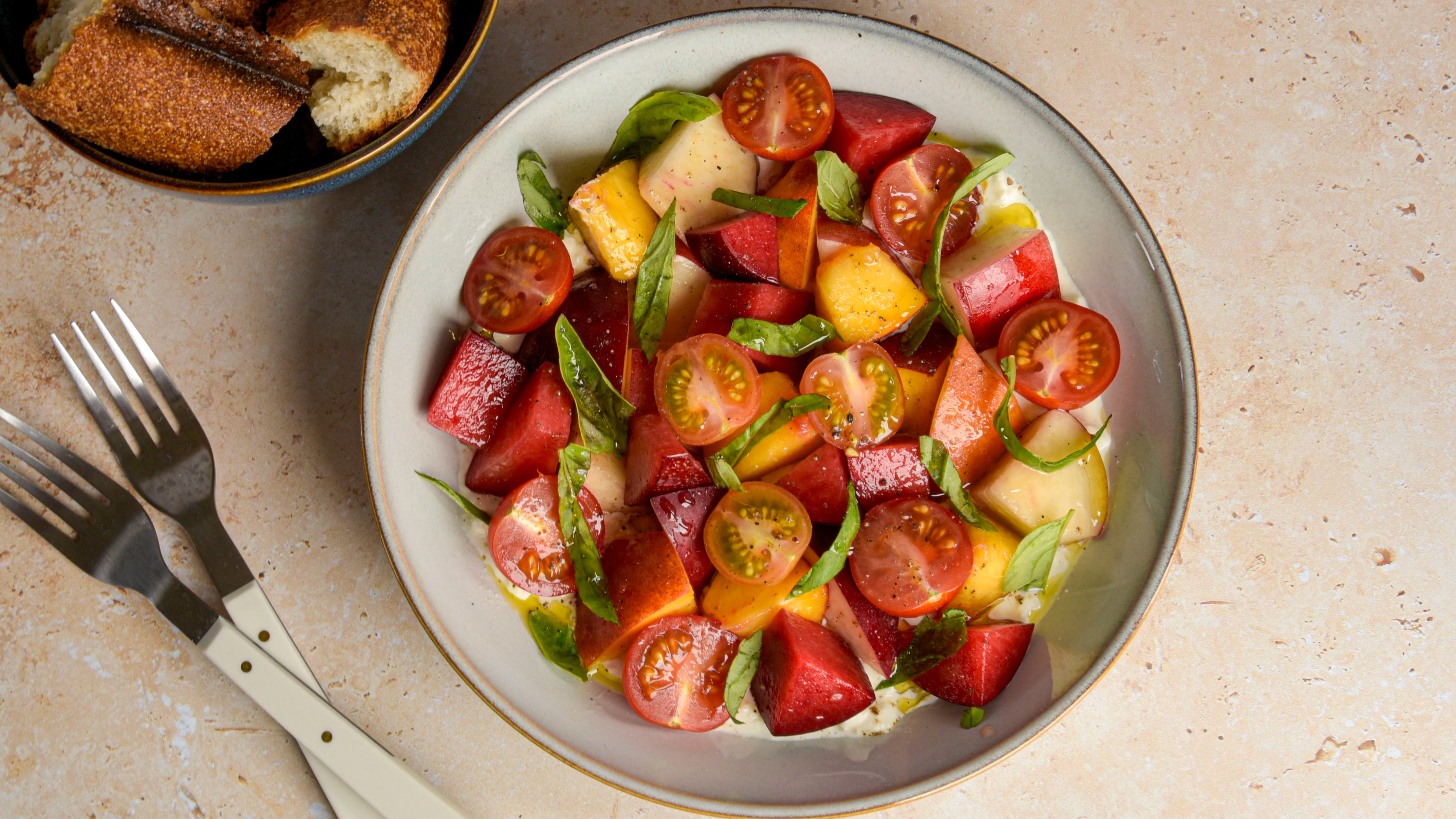 Colorful salad with tomatoes, plums, peaches and basil on a plate with bread and cutlery in the background.