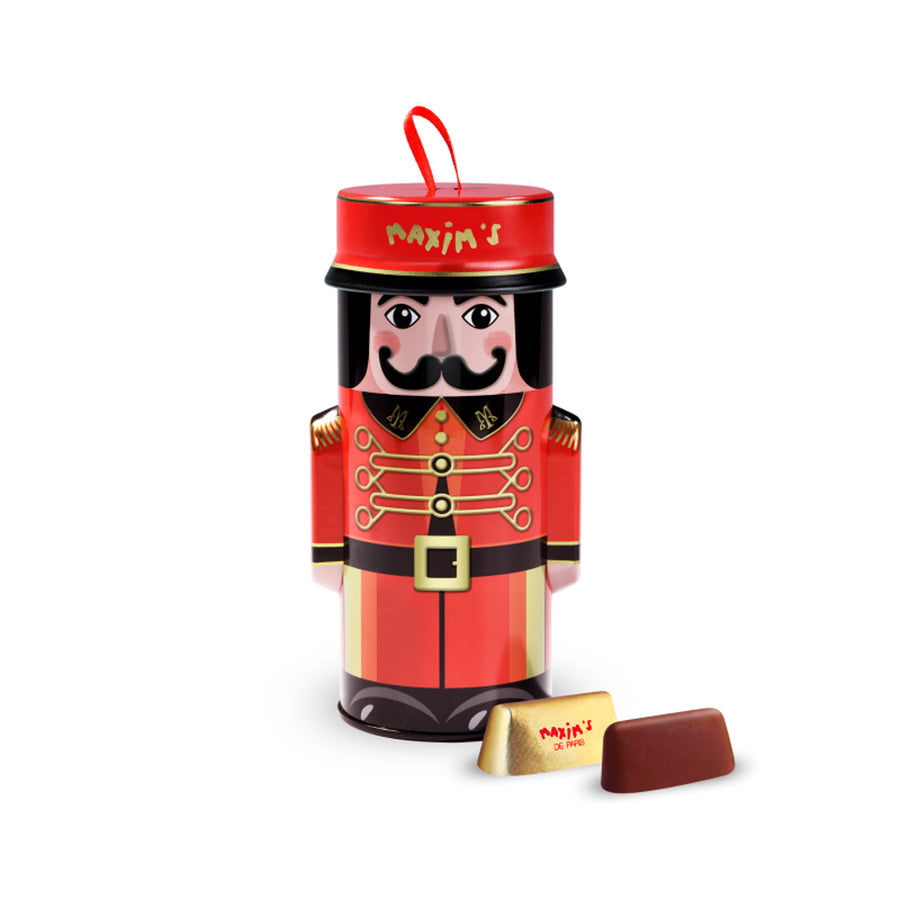 A Max's brand nutcracker-shaped chocolate tin with a red and gold uniform design, shown with a gold-wrapped chocolate and a brown chocolate piece in front, on a white background.
