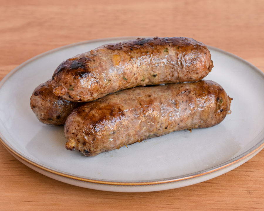Three cooked sausage links stacked on a round off-white plate with a thin gold rim, set on a light wood surface.