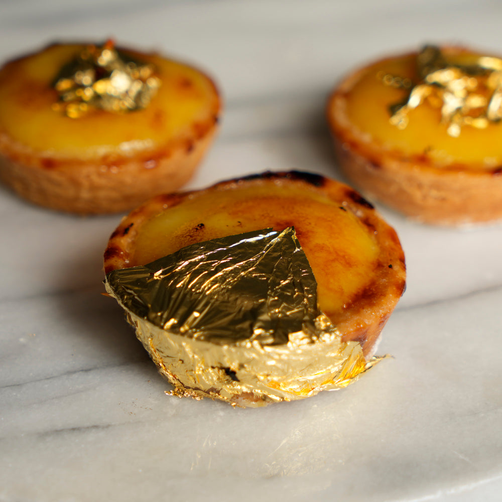Three small golden-brown tartlets with glossy custard-like fillings and leafy gold decorations, arranged on a light marble surface with shallow depth of field.