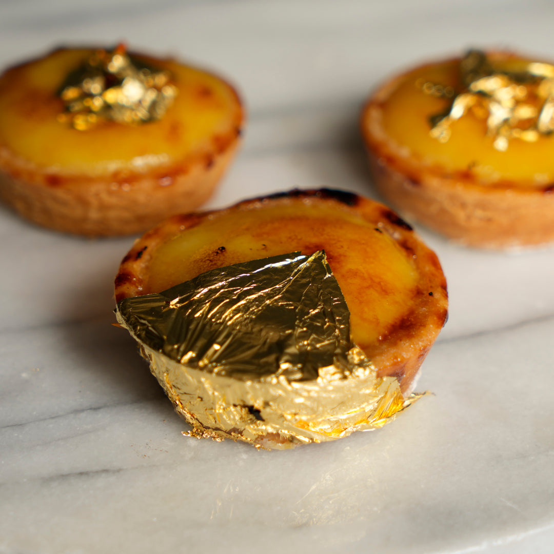 Three small golden-brown tartlets with glossy custard-like fillings and leafy gold decorations, arranged on a light marble surface with shallow depth of field.