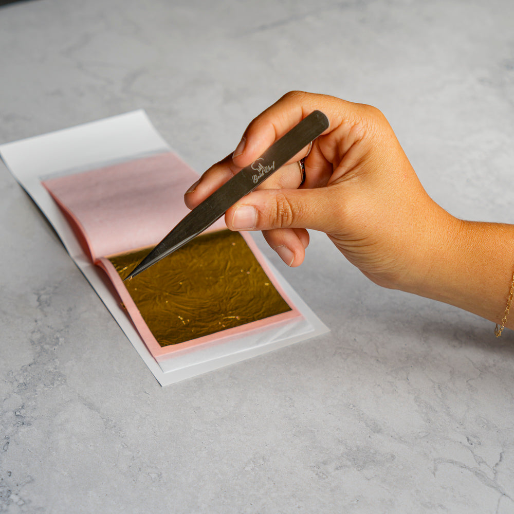 A hand holds a metal tweezers over a sheet of shiny gold leaf on pink backing paper, placed on a light gray marble surface.