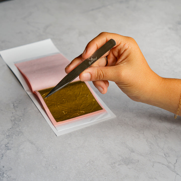 A hand holds a metal tweezers over a sheet of shiny gold leaf on pink backing paper, placed on a light gray marble surface.