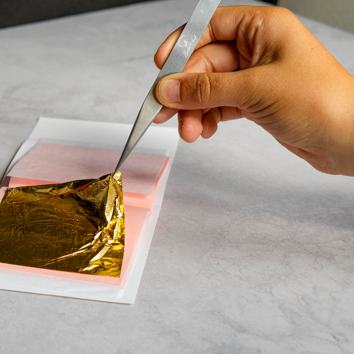 A close-up of a hand holding tweezers and lifting a shiny gold leaf sheet from a stack of translucent pink sheets on a light gray surface.